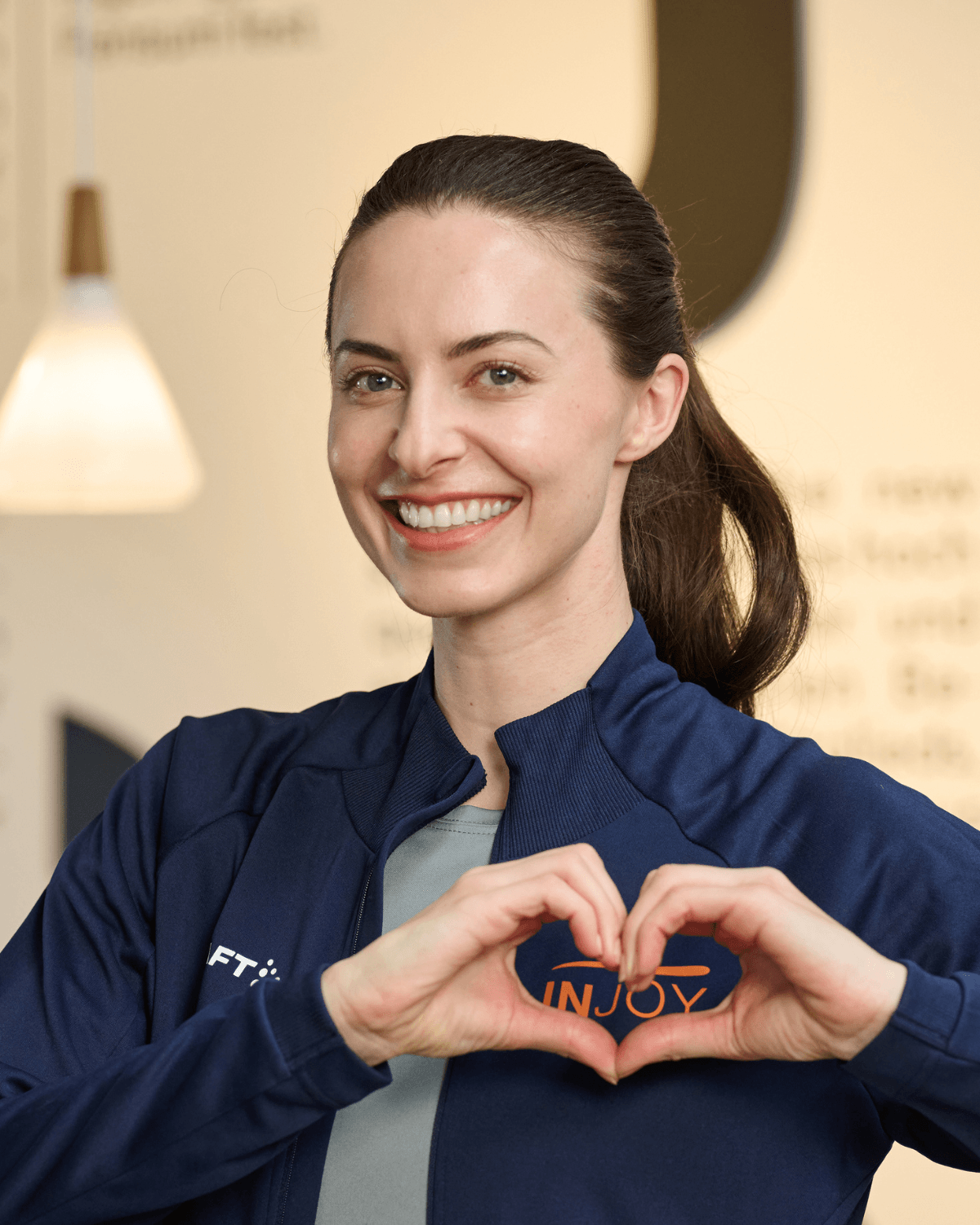 Eine INJOY Trainerin formt mir ihren Händen ein Herz, welches das INJOY Logo auf ihrer Trainingsjacke umschließt.